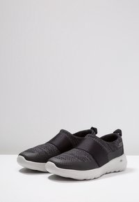 Une paire de chaussures de sport noires à enfiler avec une tige en maille texturée, un bande élastique au milieu du pied et une semelle blanche rembourrée, sur fond gris.