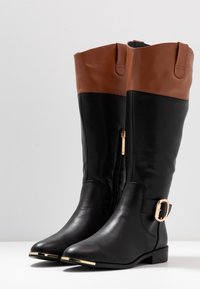 Botas altas de cuero negro con puños marrones, hebilla dorada en el lateral, cremallera dorada y acentos dorados en los dedos.