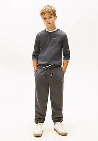 Blonder Junge steht mit den Händen in den Taschen, trägt einen dunkelgrauen Kapuzenpullover und passende Jogginghosen mit weißen Turnschuhen vor einem weißen Hintergrund.