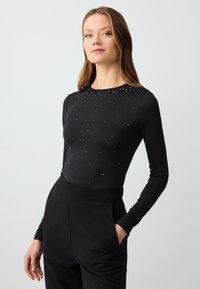 Top lungo nero a maniche, con un design aderente, caratterizzato da piccole decorazioni argento sparse nella zona del petto e un collo rotondo.