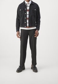 Zwarte spijkerjas, wit shirt met horizontale strepen, zwarte geplooide broek en zwarte schoenen met veters. Strakke lijnen en gestructureerde pasvorm.