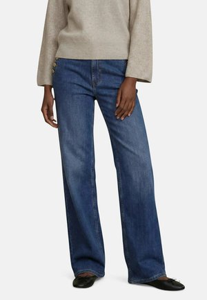 Person iført brede blå jeans med knapper i siden og en beige ribstrikket sweater, kombineret med sorte flade sko.