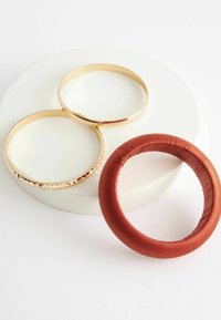 Tre armband på en vit yta: två guldmetallarmband, ett dekorerat med en strukturerad kant, plus ett rött tyginslaget armband.