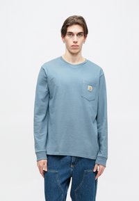 Długo rękawowa niebieska koszulka z bawełny z okrągłym dekoltem i kieszonką na piersi z kwadratową naszywką z logo. Noszona z niebieskimi dżinsami.