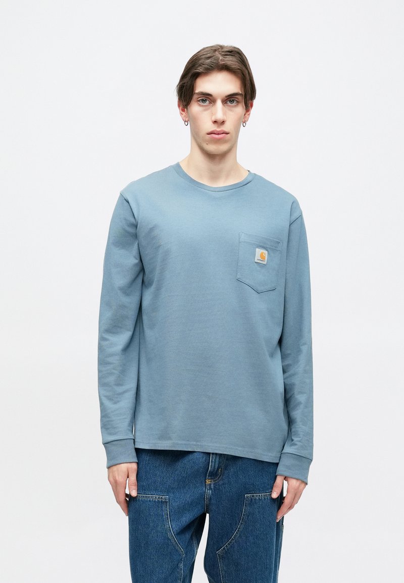 Langærmet blå bomulds t-shirt med rund halsudskæring og lomme på brystet med et firkantet logo-mærke. Bæres med blå denimbukser.