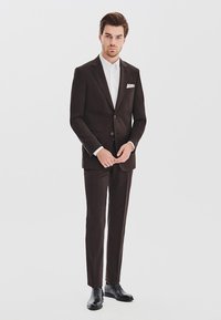 Brązowy garnitur o dopasowanym kroju z dwoma guzikami, biała koszula i poszetka. Błyszczące czarne buty dopełniają stylizację.