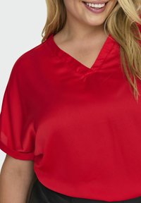 Röd kortärmad blus med V-ringning. Tillverkad av slätt tyg, med en avslappnad passform och en subtil lyster. Framhäver halsringningen.
