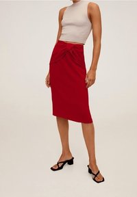 Jupe rouge ajustée avec un détail de nœud à l'avant, fabriquée en tissu lisse. Associée à un haut sans manches beige et des sandales noires à lanières.