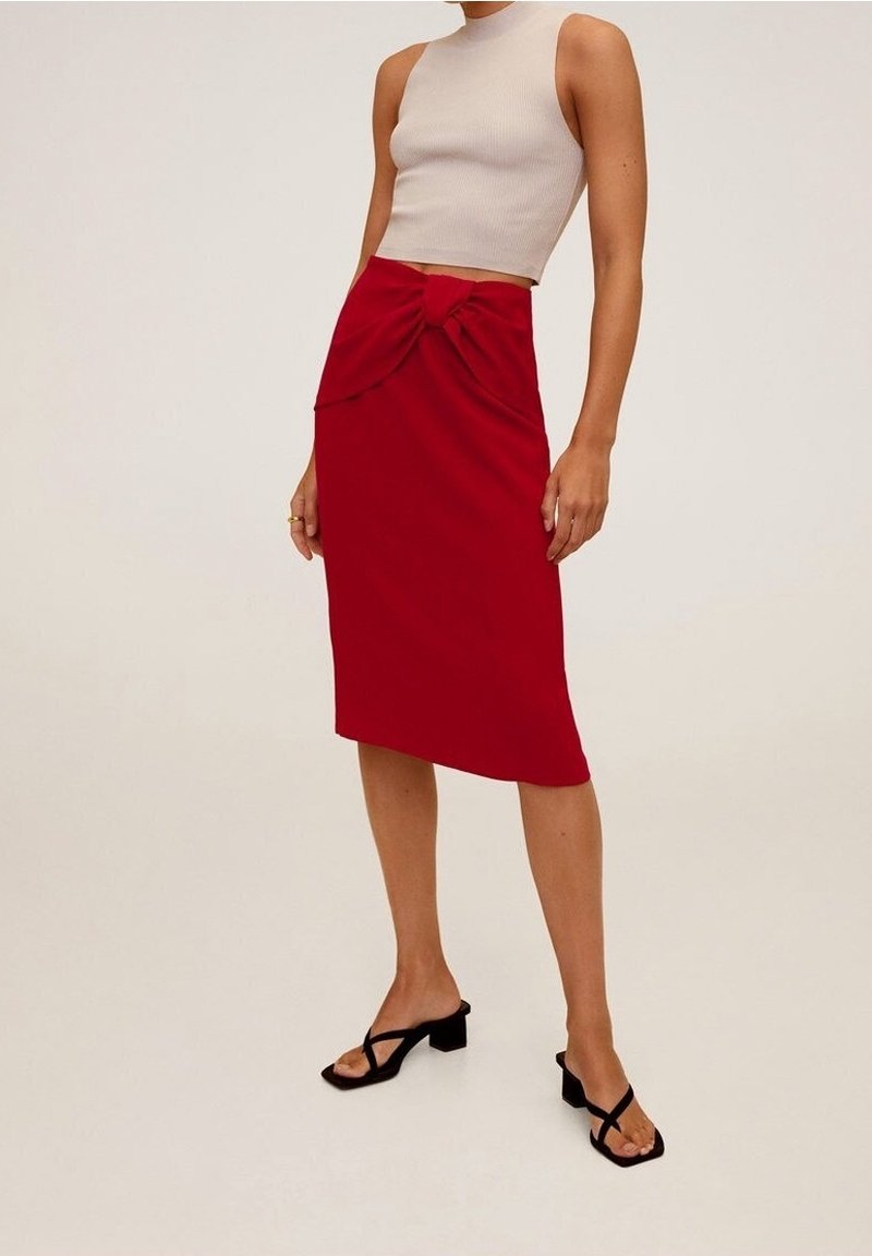 Jupe rouge ajustée avec un détail de nœud à l'avant, fabriquée en tissu lisse. Associée à un haut sans manches beige et des sandales noires à lanières.
