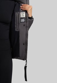 Veste rembourrée grise avec un intérieur matelassé et des boutons, arborant des étiquettes indiquant "Une étreinte chaleureuse en tissu" et le nom de la marque.