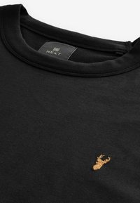 Sudadera negra con cuello redondo. Presenta un pequeño logo de ciervo dorado bordado en el lado izquierdo. Material suave y diseño sencillo.