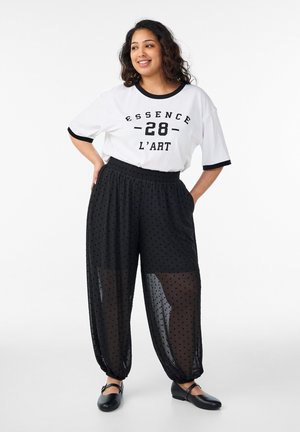 Młoda kobieta uśmiechająca się, ubrana w biały t-shirt z nadrukiem "ESSENCE 28 L'ART" oraz przezroczyste czarne spodnie w groszki, do tego czarne baleriny.