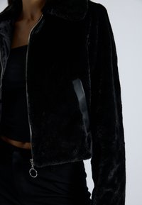 Zwarte nepbont cropped jack met een ritssluiting aan de voorkant, leren accenten en een kraag. Beschikt over een textuurafwerking en een ronde ritstrekker.