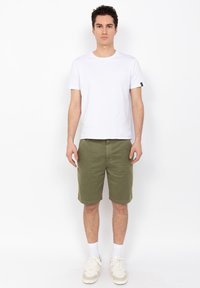 Camiseta blanca de manga corta combinada con pantalones cortos verde oliva. El modelo lleva zapatillas blancas con detalles en marrón y calcetines blancos, de pie contra un fondo sencillo.