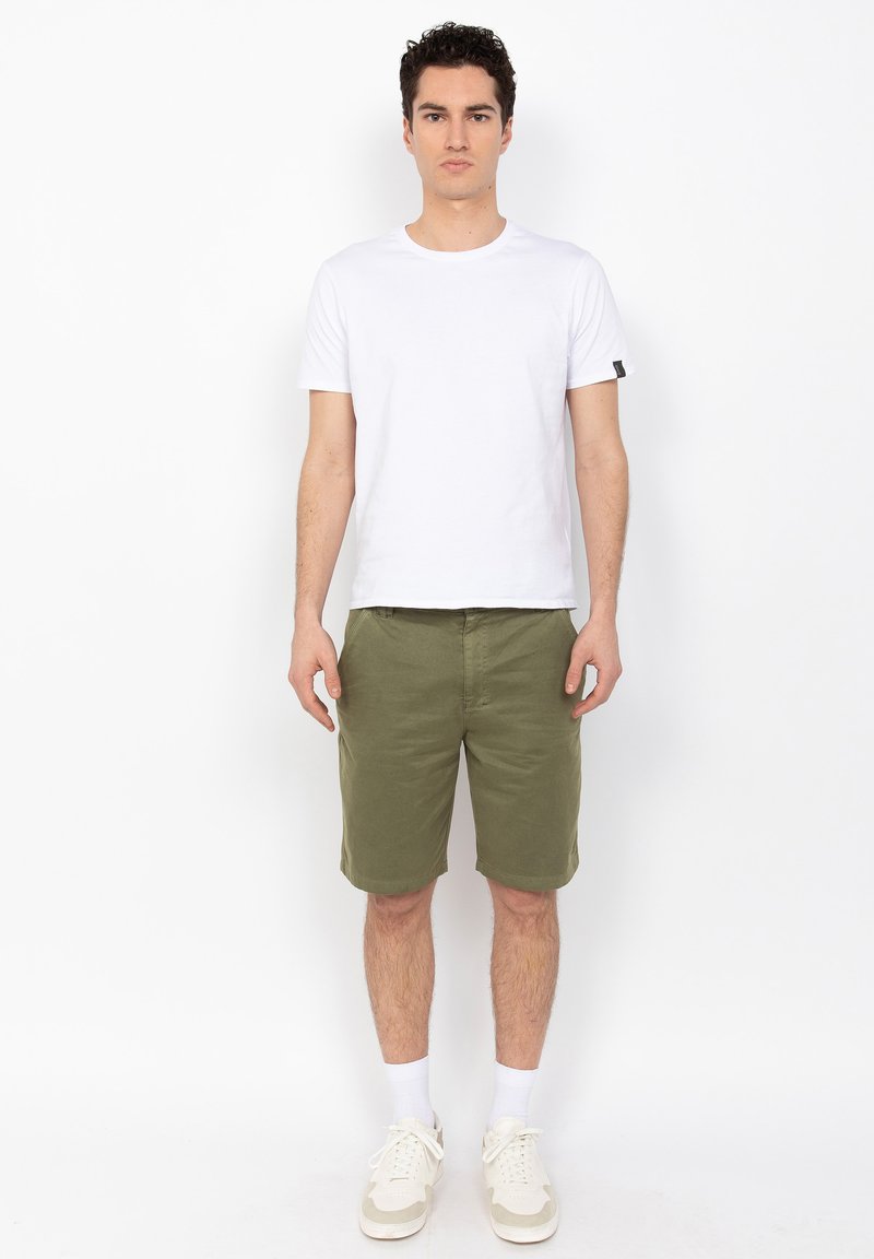 Camiseta blanca de manga corta combinada con pantalones cortos verde oliva. El modelo lleva zapatillas blancas con detalles en marrón y calcetines blancos, de pie contra un fondo sencillo.