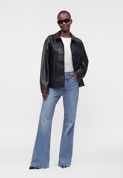 Personne portant des lunettes de soleil, une veste en cuir noire, une chemise blanche et un jean bleu évasé, se tient avec assurance devant un fond uni.