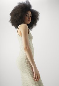 Κρεμ φορεμάτα, εφαρμοστό με υφή. Διαθέτει αμάνικο σχέδιο που φτάνει μέχρι την μέση της γάμπας. Λείος τελείωμα και διακριτική γυαλάδα.