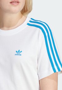 Vit bomull t-shirt med blå tre-randig design på ärmarna och blå broderad Adidas-logotyp på bröstet, rundad halsringning, korta ärmar.