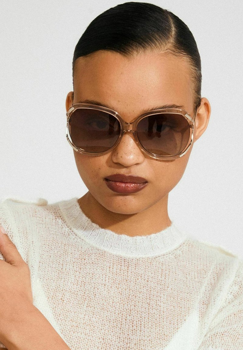 Hnědé nadměrné sluneční brýle s průhledným béžovým rámem a tmavými čočkami. Mají kulatý tvar a elegantní nožičky. Nošené s lehkým texturovaným topem.