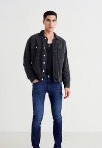 Joven de pie contra un fondo blanco, vestido con una chaqueta de mezclilla negra, una camisa negra, jeans azules y un collar con un colgante.