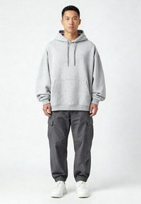 Felpa grigia con cappuccio e tasca frontale, abbinata a pantaloni cargo grigi scuri con tasche laterali. Le scarpe sono sneakers bianche.