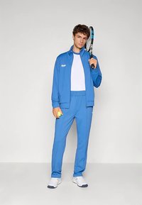 Sinine sportlik dress, millel on lukuga jakk ja elastse vöökohaga püksid, kantud valge särgi peal. Hoides käsis tennise reketit ja palli.