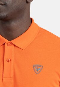 Oranžové polo tričko s texturovanou tkaninou, vybavené límečkem, dvěma odpovídajícími knoflíky a modrou nášivkou na levém hrudníku.