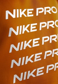 Texte blanc répété "NIKE PRO" imprimé sur un fond de tissu orange, incliné en diagonale sur la surface.