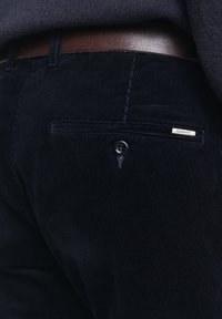 Pantalon en velours côtelé bleu foncé avec des rayures verticales, doté d'une poche arrière avec un bouton et d'une étiquette de marque. Le matériau semble doux et texturé.
