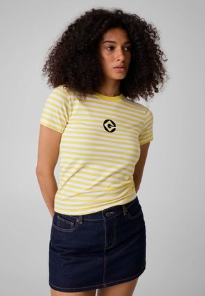 Femme aux cheveux bouclés portant un t-shirt rayé jaune et blanc avec un logo noir et une jupe en denim foncé, debout devant un fond gris.