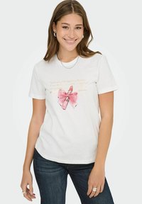 Jonge vrouw in een wit T-shirt met een roze lippenstift en strikgraphic, blauwe jeans, een zilveren ketting en oorbellen met een ring, die naar de camera lacht.