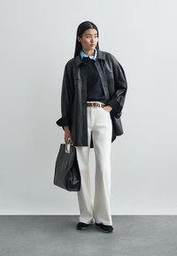 Chemise oversize en cuir noir, pull bleu marine, pantalon blanc, ceinture marron, grand sac fourre-tout noir, baskets noires, chemise à col légère bleu clair en dessous.