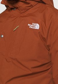 Hnědá nepromokavá bunda s přední kapsou, elastickým stahovacím kapucí a bílým vyšívaným logem na hrudi. Hladká textura.