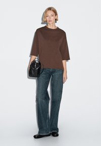 Woman wearing brown oversized top and wide-leg jeans, holding a small black leather backpack, standing against a plain white background.