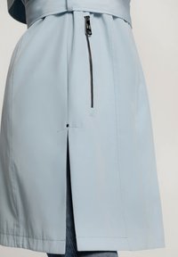 Lichtblauwe jas met een voorzak met rits, getailleerd ontwerp en een zijsplit. Gladde textuur met precieze stikdetails.