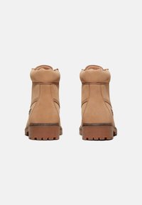 Hnědé semišové kotníkové boty s kulatou špičkou, ohrnutým límcem a texturovanou gumovou podrážkou. Mají minimální švy a dekorativní boční prvek.