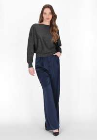 Schwarzes, glitzerndes Langarmoberteil mit weitem Ausschnitt, kombiniert mit navyblauen, weit geschnittenen Hosen aus glattem Material und engen Bündchen. Schwarze High Heels runden das Outfit ab.