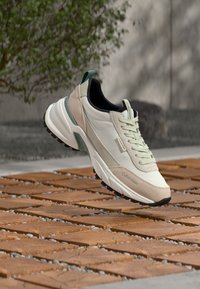 Αθλητικό παπούτσι Calvin Klein σε ανοιχτό μπεζ και λευκό με απαλές πράσινες λεπτομέρειες, αιωρούμενο πάνω από ξύλινο δάπεδο με πλακάκια και θολό εξωτερικό φόντο.