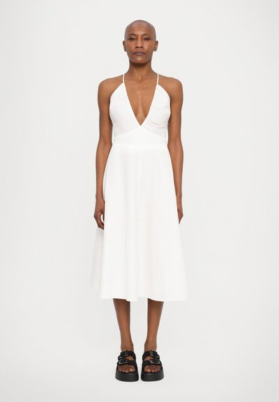 White A-line dress with a deep V-neck and thin straps, featuring a flowing skirt. Black platform sandals with straps complete the look.