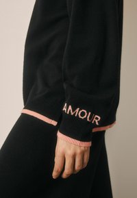 Črna majica s pestro obrobo na rokavih v rožnati barvi, na levem rokavu je izvezeno besedo "AMOUR". Vidna mehka pletena tekstura.