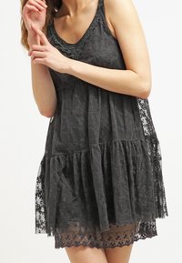 Robe en dentelle noire avec un design à étages, un ourlet festonné et des motifs floraux. Tissu léger et transparent avec un corsage ajusté et des bretelles larges.