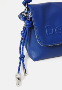 Borsa a tracolla in pelle blu royal con logo impresso, chiusura a zip e manici decorativi intrecciati con hardware in tonalità argento.