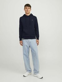 Marineblaue Kapuzenpullover mit Fronttasche, Kordelzügen und Logodetail; kombiniert mit hellblauen, locker sitzenden Jeans und weißen Sneakers.