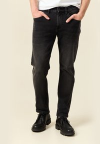 Hombre vestido con jeans negros y zapatos de cuero negros con cordones, con las manos en los bolsillos, de pie contra un fondo beige.