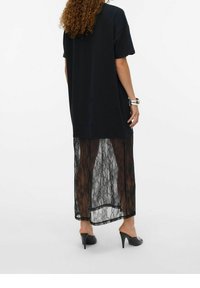 Nainen, jolla on yllään musta ylisuuri t-paitamekko, jonka alaosa on läpinäkyvää pitsiä, mustat korokepohjaiset sandaletit ja hopea rannekoru, kääntyy poispäin.