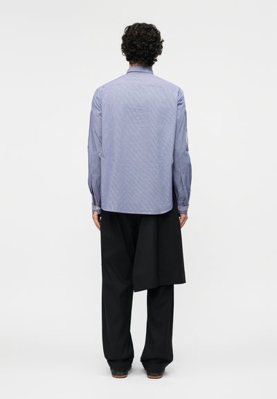 Homme aux cheveux bouclés portant une chemise bleu clair à manches longues et un pantalon noir à jambes larges, debout de dos sur un fond uni.