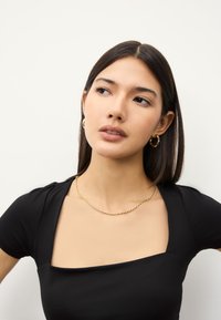 Schwarzes, figurbetontes Oberteil mit quadratischem Ausschnitt, strukturiertem Stoff und kurzen Ärmeln. Goldene Halskette mit Kettengliedern und Creolen runden den Look ab.