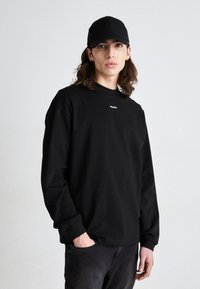 Black long-sleeve sweatshirt with a crew neck and white "HUGO" logo on the chest, paired with a black cap. Casual design, minimalistic style.