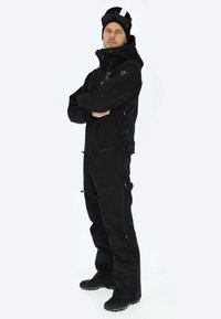 Salopette nera con zip e tasche, chiusura regolabile e cappuccio aderente. Materiale testurizzato e impermeabile, progettata per sport invernali.