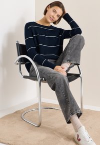 Navy striped knit sweater with white lines, paired with gray jeans. Model seated on a chrome chair with textured beige rug underneath.
